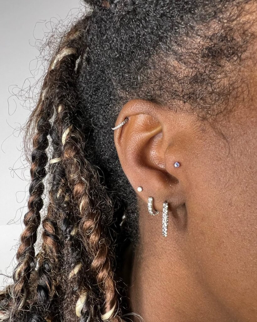 Oreja decorada con múltiples piercings, incluyendo una herradura en el cartílago superior, un lóbulo secundario con una joya púrpura y dos pendientes largos de cristal colgando del lóbulo.