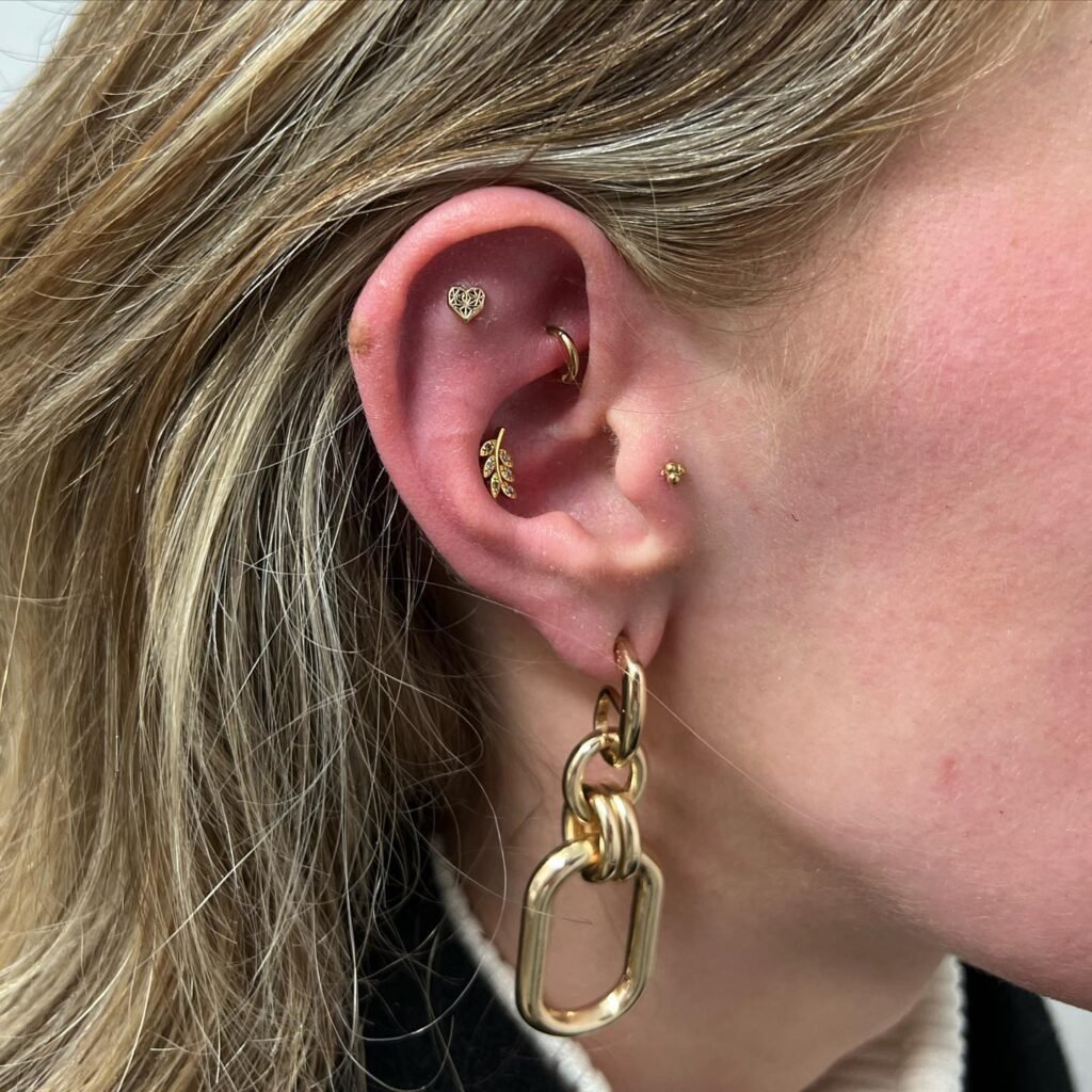 ista lateral de una oreja adornada con varios piercings de oro, incluyendo un pequeño corazón en el cartílago superior, una pieza en forma de hoja en el tragus y un aro dorado en el lóbulo junto a un pendiente de gran eslabón colgante.